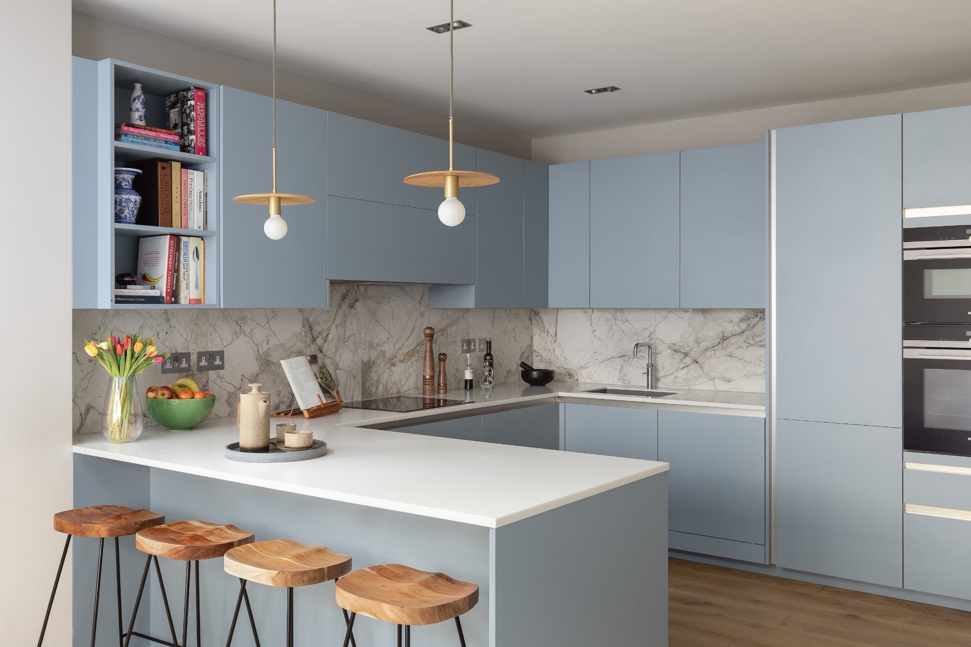 Pale blue kitchen peninsula with marble-effect splashback, timber bar stools and brass pendant lights