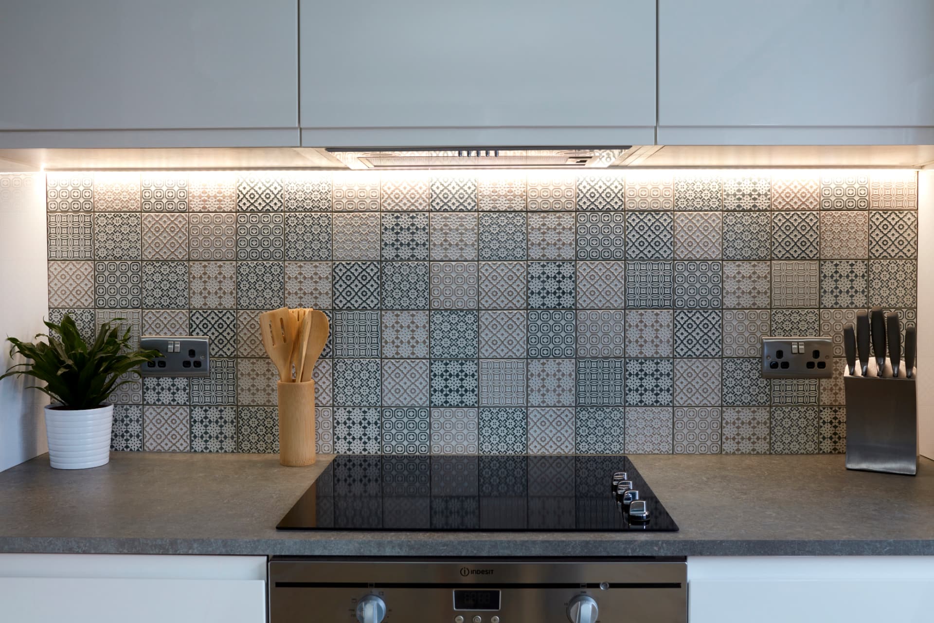 Patterned grey and white splashback tiles behind an induction hob, under-cabinet lighting