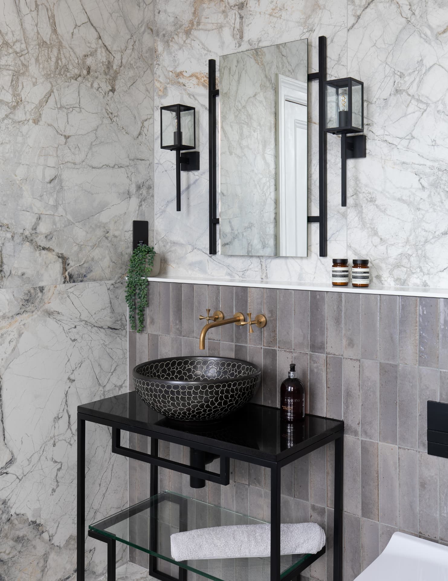Bathroom vanity with a patterned black vessel basin, wall-mounted brass tap, ribbed grey tile splashback, mirror and black wall lights