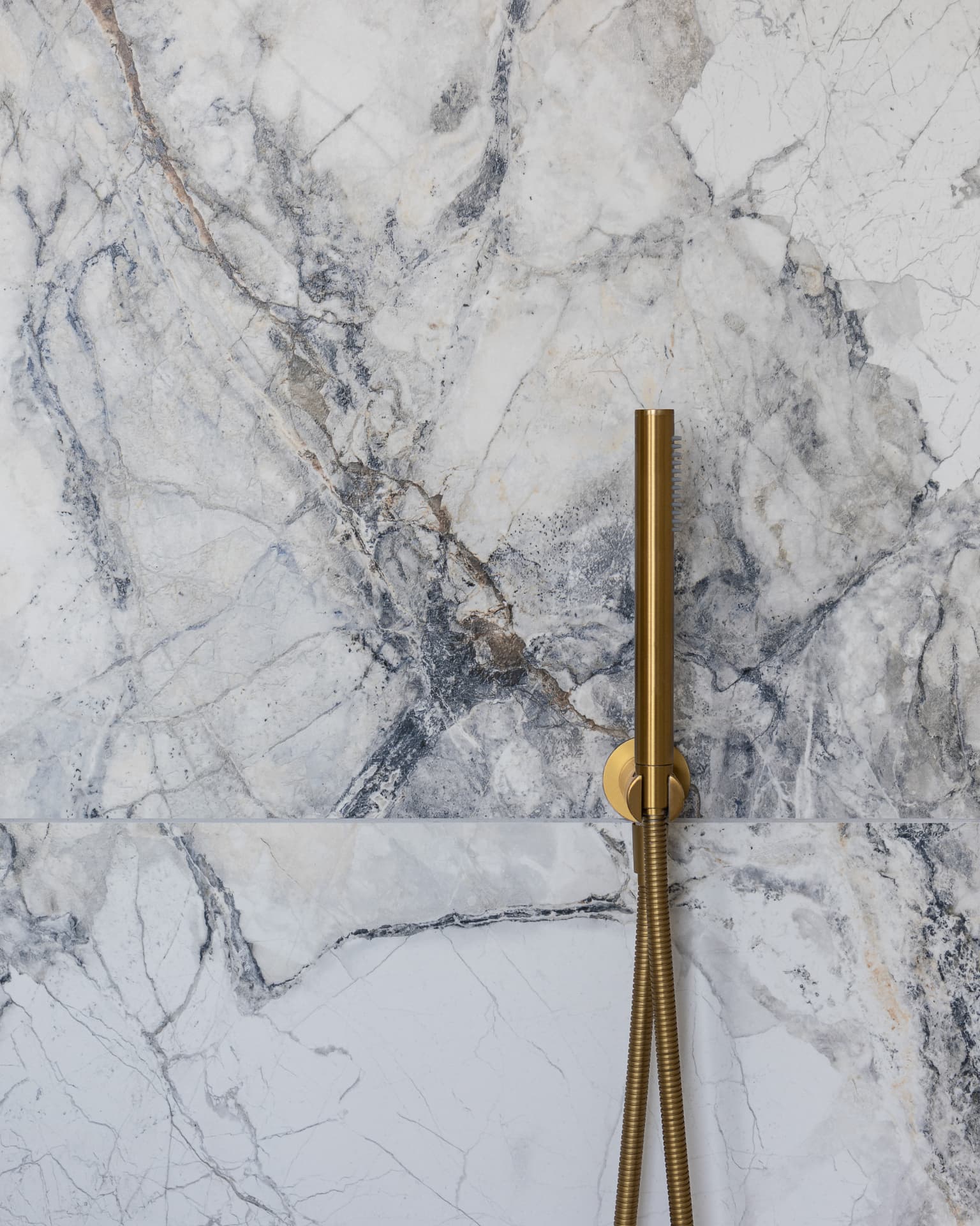 Brushed brass handheld shower wand and hose against marble-effect wall tiles