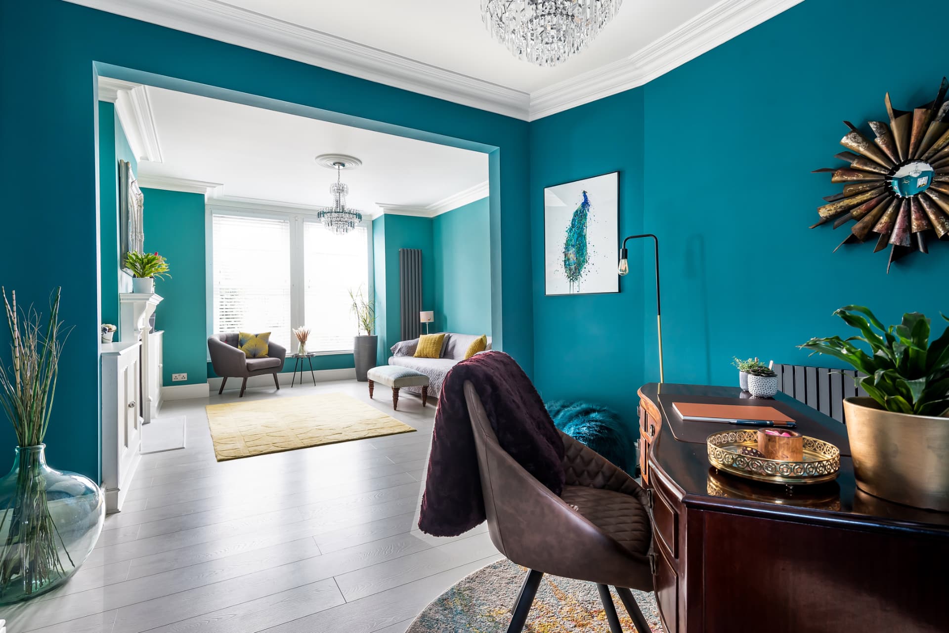 Open-plan living and dining room in teal with bay window, peacock feather mirror and writing desk