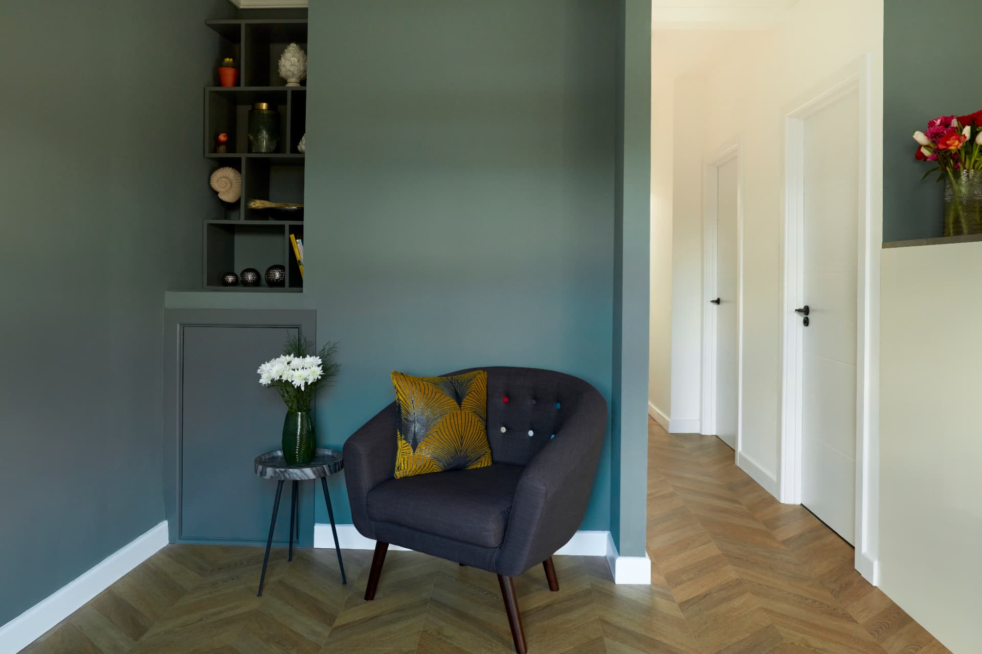 Reading corner with grey armchair and built-in display niche, beside a hallway with herringbone floor