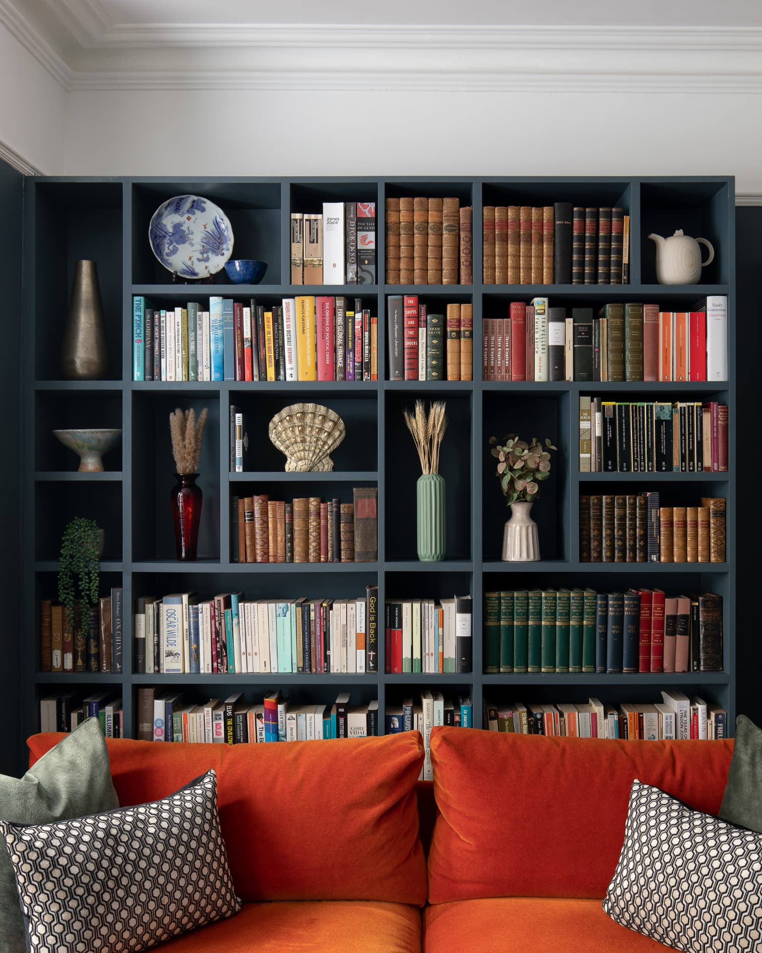Floor-to-ceiling bespoke library shelving painted dark blue above an orange velvet sofa