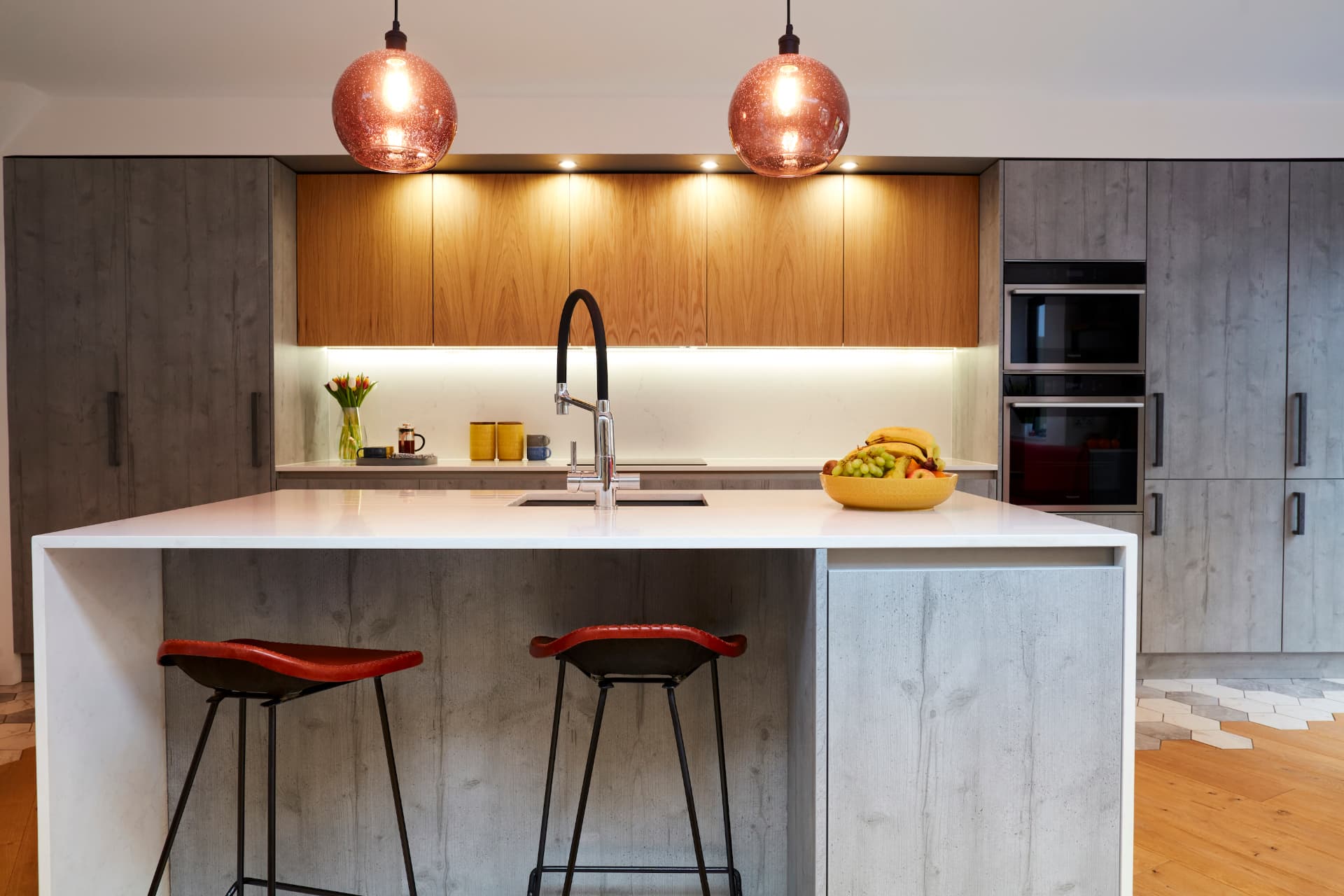 Kitchen island with white waterfall worktop, red bar stools and copper pendant lights in front of walnut wall units