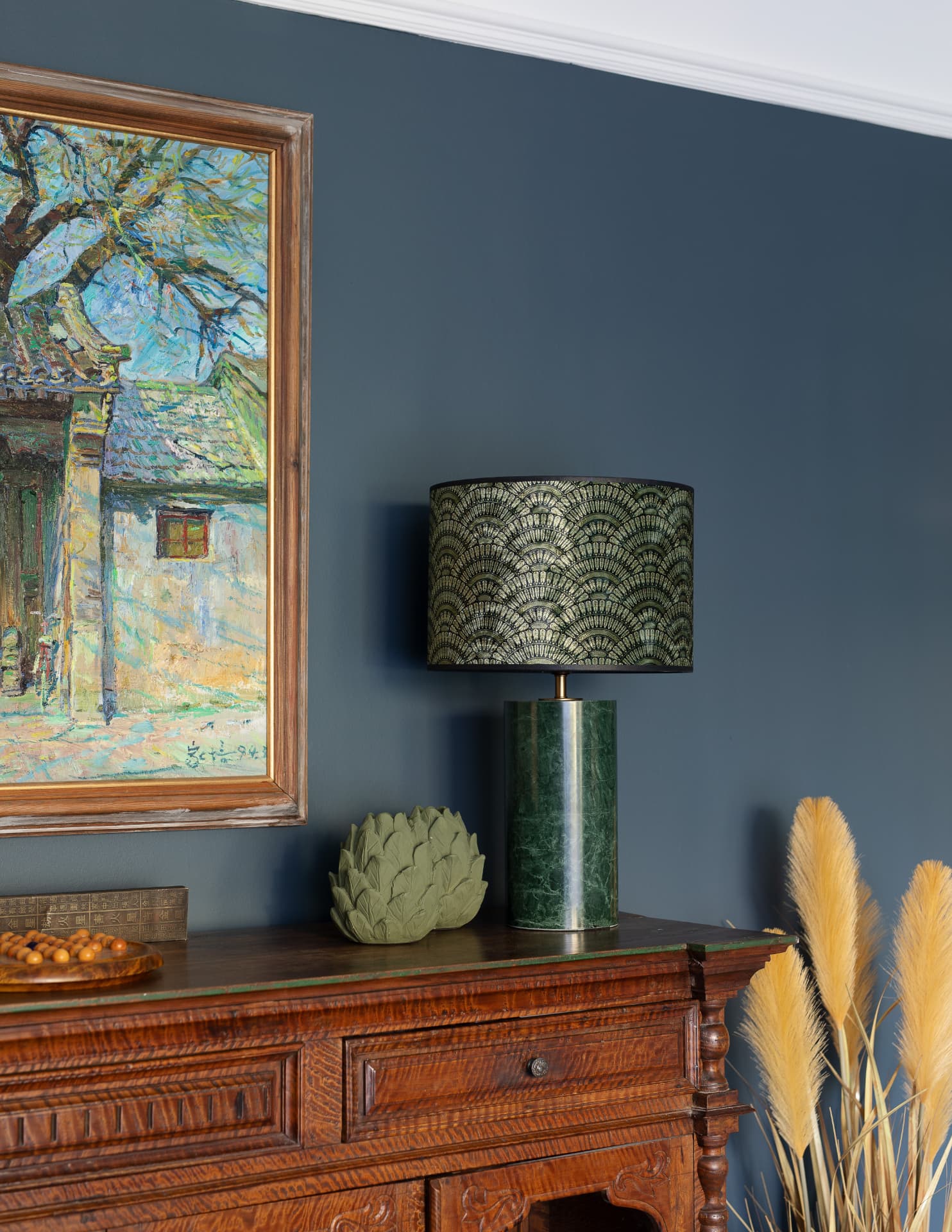 Antique carved sideboard with green ceramic lamp and framed oil painting against a deep blue wall