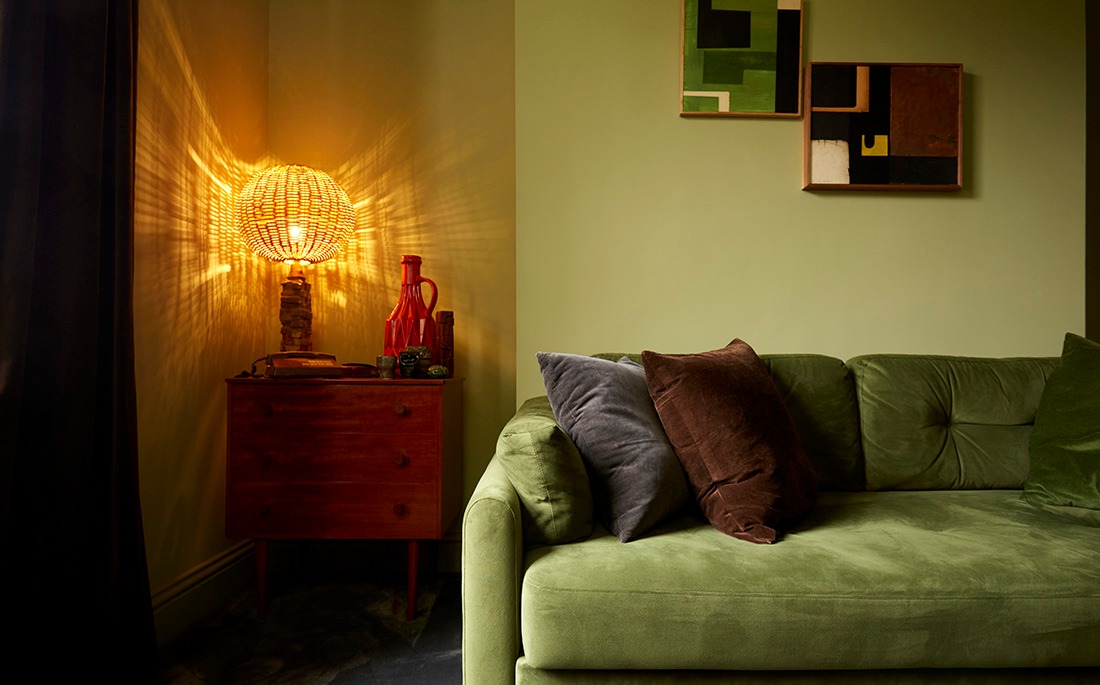 Living room corner in a Hackney renovation with green velvet sofa, teak sideboard and glowing table lamp