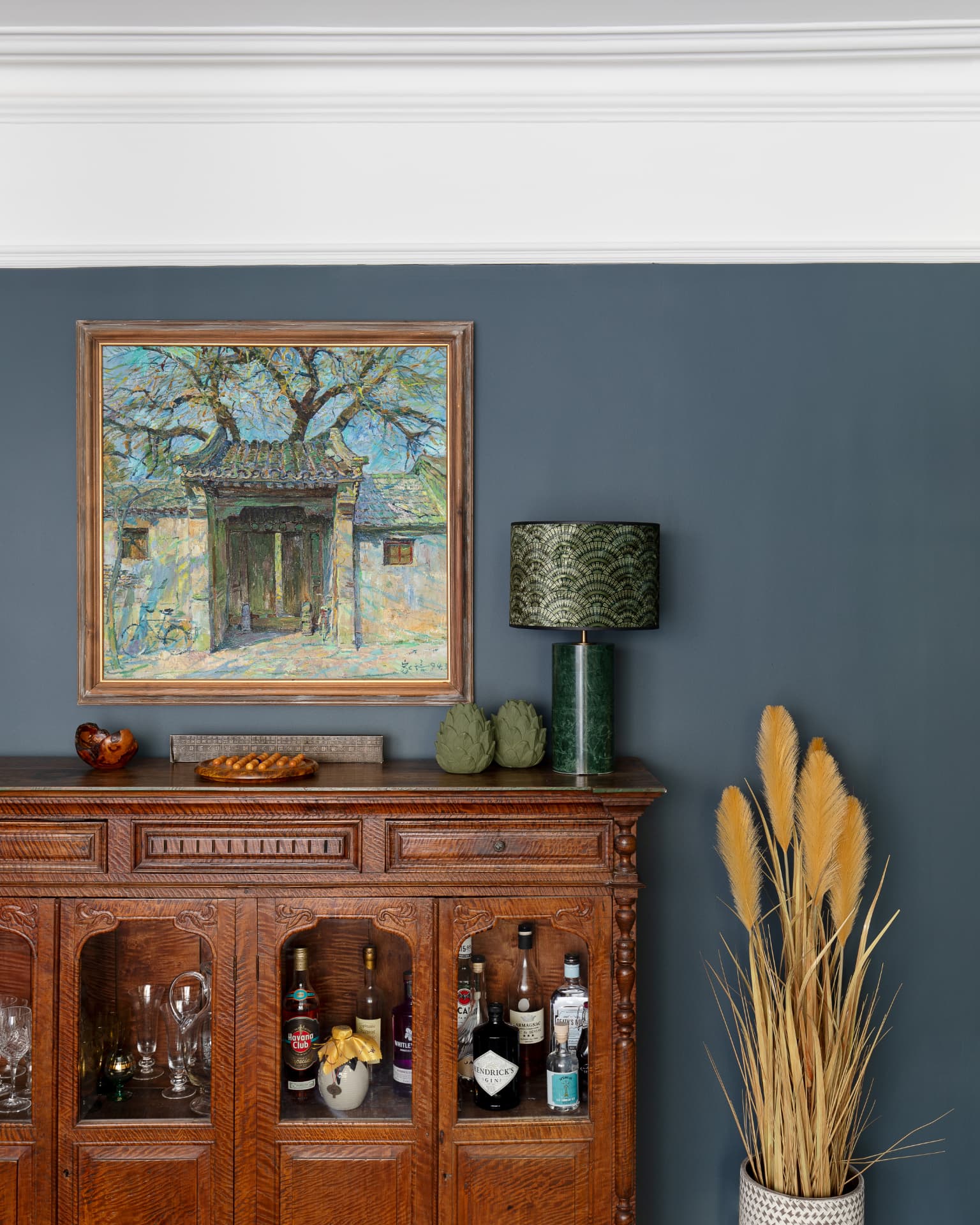 Carved wooden drinks cabinet beneath a framed painting on a deep blue living room wall